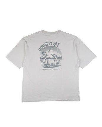 La camiseta VAQUERO VAQUERA Houston es una camiseta unisex de color gris claro con un gráfico en la espalda de un astronauta y un tiburón en el espacio, con "HOUSTON" encima y "TENEMOS UN PROBLEMA" debajo, una atrevida mezcla de estilo occidental y estilo cósmico.