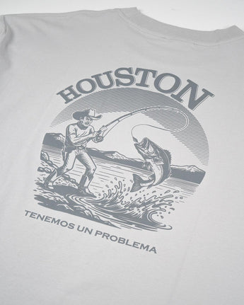 La Houston Tee de VAQUERO VAQUERA es una playera gris unisex con ilustración western de vaquero pescando, montañas y texto: HOUSTON arriba y TENEMOS UN PROBLEMA abajo.