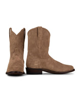 La VAQUERO VAQUERA Alamo Bota de Gamuza Bentley es una bota western de ante color tostado con correas de tiro y tacón bajo de madera, mostrada con una bota erguida y la otra inclinada hacia delante.