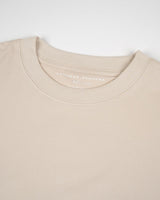 Un primer plano de la camiseta Beige Tee de VAQUERO VAQUERA, colocada en horizontal para mostrar el escote, los detalles de costura y la zona superior del pecho. La etiqueta interior muestra el nombre de la marca y la talla M.