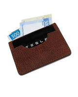 Tarjetero De Res VAQUERO VAQUERA de diseño minimalista en color marrón, con una tarjeta negra TESLA y dos billetes de 500 pesos parcialmente visibles, aislado sobre fondo blanco.