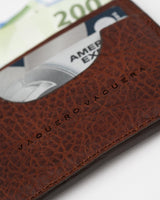 Un primer plano del Tarjetero De Res de VAQUERO VAQUERA, con "VAQUERO VAQUERA" en relieve, muestra un billete de 200 euros y una tarjeta American Express parcialmente visibles en su diseño minimalista de cuero marrón.
