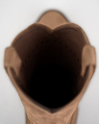 Vista de arriba abajo de la apertura de un Alamo Bota de Gamuza Bentley color canela de VAQUERO VAQUERA, con "VAQUERO" impreso en el forro interior oscuro. El fondo es de color claro y está desenfocado.