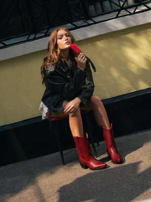 Una joven con chaqueta negra y Santa Bárbara Bota De Res Roja de VAQUERO VAQUERA está sentada al aire libre en una silla, lamiendo un polo rojo. Está delante de una pared amarilla con una ventana encima.