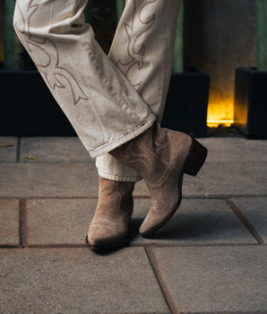 Una persona con pantalones bordados de color claro y VAQUERO VAQUERA Santa María - Bota de Gamuza Shell de pie sobre un pavimento de piedra con las piernas cruzadas; iluminación amarilla suave y una pared verdosa en el fondo.