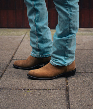 Una persona vestida con vaqueros azul claro y VAQUERO VAQUERA Murrieta - Whisky Bota de Gamuza se encuentra de pie en una acera asfaltada, con sólo la parte inferior de las piernas y los pies visibles.