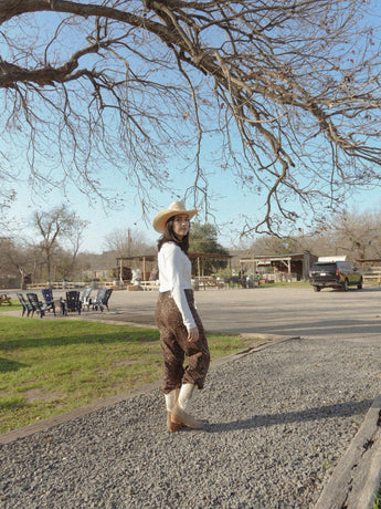 Con sombrero vaquero, top blanco, pantalones estampados y botas Santa Sofía Bota De Res y Gamuza Beige de VAQUERO VAQUERA, una persona se encuentra en un camino de grava al aire libre cerca de hierba y edificios de un rancho con un coche al fondo.