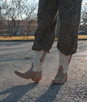 Una persona con pantalones de estampado de leopardo y Santa Sofía - Bota de Gamuza Crudo de VAQUERO VAQUERA con dibujos florales está de pie en una acera iluminada por el sol, proyectando largas sombras, con árboles y una valla de madera al fondo.