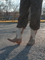 Una persona vestida con pantalones marrones estampados y VAQUERO VAQUERA Santa Sofía Bota De Res y Gamuza Beige con un diseño floral está de pie al aire libre sobre una superficie pavimentada, con árboles y una valla visibles en el fondo.