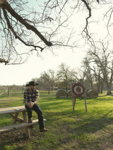 Un hombre con camisa de cuadros y sombrero vaquero, que lleva la VAQUERO VAQUERA Arrieta Bota de Cierre Pitón Canela, se sienta en una mesa de picnic de madera en un campo cubierto de hierba con balas de heno y una diana de tiro con arco bajo un cielo parcialmente nublado.