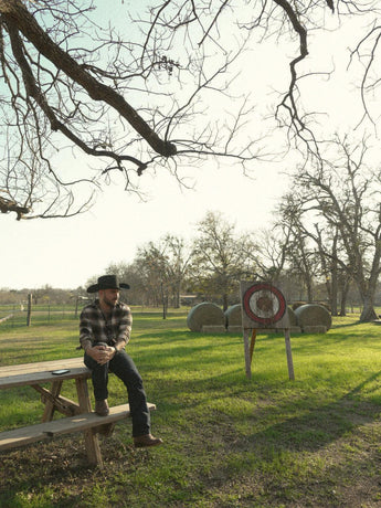 Un hombre con camisa de cuadros y sombrero vaquero, que lleva la VAQUERO VAQUERA Arrieta Bota de Cierre Pitón Canela, se sienta en una mesa de picnic de madera en un campo cubierto de hierba con balas de heno y una diana de tiro con arco bajo un cielo parcialmente nublado.