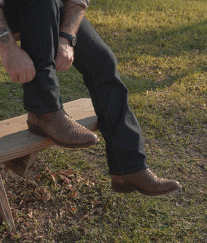 Una persona con vaqueros oscuros y Arrieta - Bota de Cierre Pitón Nogal de VAQUERO VAQUERA se relaja en un banco de madera al aire libre, con la hierba y la luz del sol de fondo.