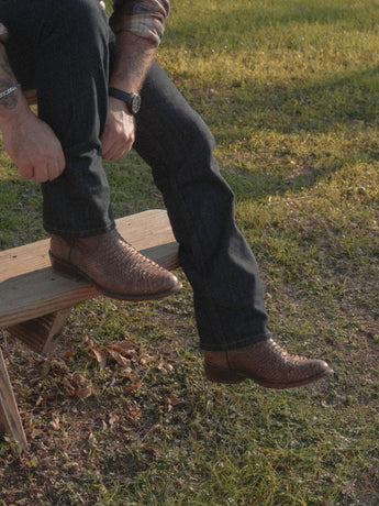 Una persona vestida con vaqueros y la exclusiva VAQUERO VAQUERA Arrieta Bota de Cierre Pitón Canela está sentada en un banco de madera al aire libre, rodeada de hierba y hojas caídas. Sólo se ve la parte inferior de su cuerpo.