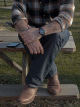 Una persona sentada en una mesa de picnic de madera, con las piernas cruzadas, lleva vaqueros, una camisa de cuadros, una Arrieta Bota de Cierre Pitón Canela de VAQUERO VAQUERA y un reloj de pulsera. Un smartphone está en el banco de al lado con hierba al fondo.