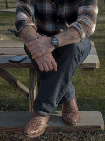 Una persona sentada en una mesa de picnic de madera, con las piernas cruzadas, lleva vaqueros, una camisa de cuadros, una Arrieta Bota de Cierre Pitón Canela de VAQUERO VAQUERA y un reloj de pulsera. Un smartphone está en el banco de al lado con hierba al fondo.