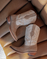 Un par de botas Santa Sofía - Bota de Gamuza Crudo de VAQUERO VAQUERA, en ante marrón claro con pespuntes decorativos, se exhiben una al lado de la otra sobre un banco de cuero acolchado color canela.