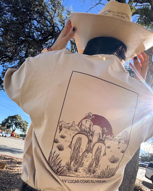 Una persona con sombrero vaquero y la camiseta La familia Tee de VAQUERO VAQUERA-una camiseta beige con dos jinetes en un desierto bajo un sol rojo-está al aire libre, con la luz del sol brillando y árboles alrededor. Destaca su estilo auténtico.