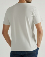 Un hombre de pie, de espaldas a la cámara, vestido con una camiseta VAQUERO VAQUERA Gris claro y unos vaqueros azul oscuro con las manos en las caderas sobre un fondo claro.