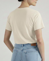 Se ve a una mujer de espaldas con una camiseta VAQUERO VAQUERA beige y unos vaqueros azul claro de cintura alta. Lleva el pelo recogido y está de pie sobre un fondo claro liso, captando un estilo vaquero desenfadado.