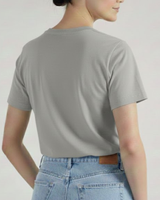 Se muestra a una persona de espaldas con una camiseta VAQUERO VAQUERA Gris claro y unos vaqueros azules de cintura alta, de pie sobre un fondo liso.