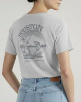 Una mujer de pie de lado, con una mano en el bolsillo de los vaqueros, lleva la camiseta VAQUERO VAQUERA Houston Tee, una camiseta unisex de color gris claro con una ilustración de pesca, "HOUSTON" y "TENEMOS UN PROBLEMA" en la espalda.