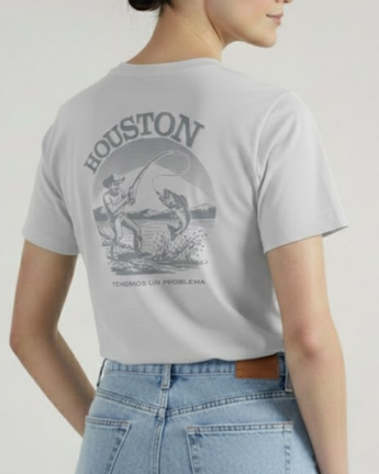 Una mujer de pie de lado, con una mano en el bolsillo de los vaqueros, lleva la camiseta VAQUERO VAQUERA Houston Tee, una camiseta unisex de color gris claro con una ilustración de pesca, "HOUSTON" y "TENEMOS UN PROBLEMA" en la espalda.