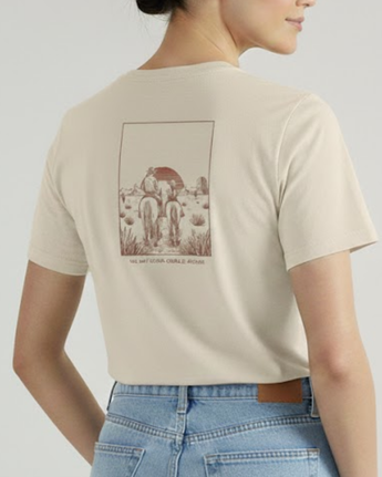 Una mujer viste La familia Tee de VAQUERO VAQUERA, una vaquera playera beige con un dibujo de tres caballos en un desierto. Ligeramente girada, luce el diseño western, combinado con unos vaqueros azules de cintura alta.