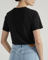 Una persona mira de espaldas a la cámara, con la camiseta Negro Tee de VAQUERO VAQUERA y unos vaqueros de cintura alta azul claro sobre un fondo liso y neutro.