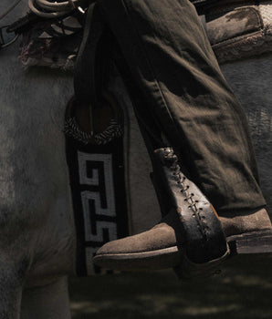Un primer plano muestra a una persona con pantalones oscuros que lleva VAQUERO VAQUERA Murrieta - Bota de Gamuza Taupe, sentada en un caballo con el pie en un estribo con un dibujo geométrico en blanco y negro.