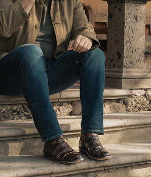 Una persona con vaqueros azules, chaqueta verde y llamativas botas VAQUERO VAQUERA Murrieta Caoba Caimán Americano está sentada en unos escalones de piedra con un brazo apoyado en la rodilla. Su rostro no es visible en la imagen.